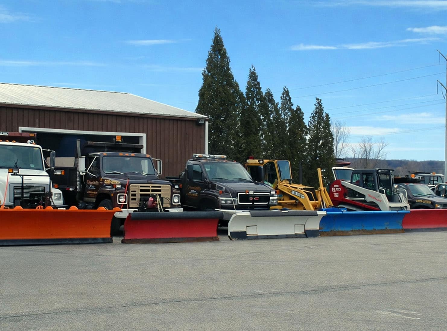5 snow plow trucks parked in a row beside each other in a lot with a large storage barn in the background